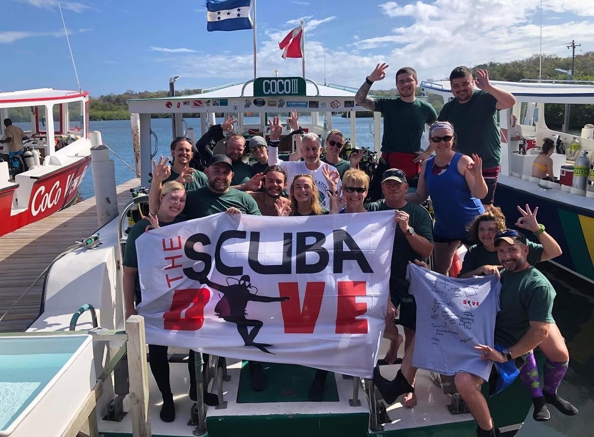 A group of divers poses on a boat holding a banner for "THE SCUBA LOVE," celebrating their diving experience in Honduras.