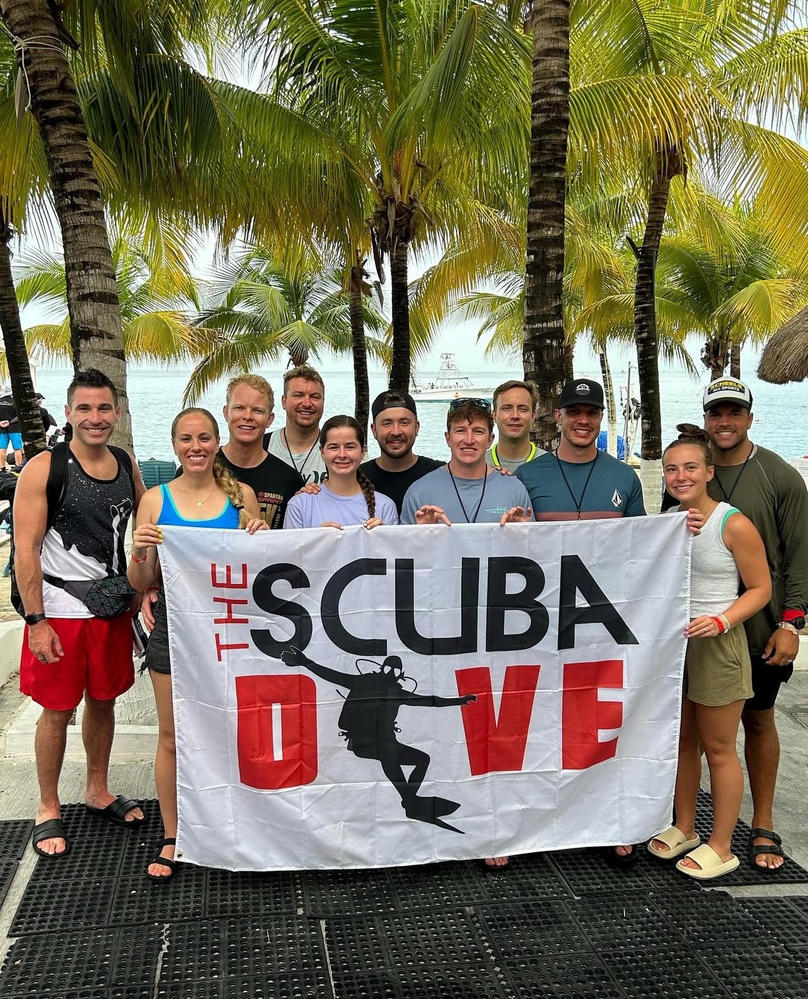 A group of ten divers stands together, holding a banner for "THE SCUBA DIVE" in a tropical setting with palm trees and a boat in the background.