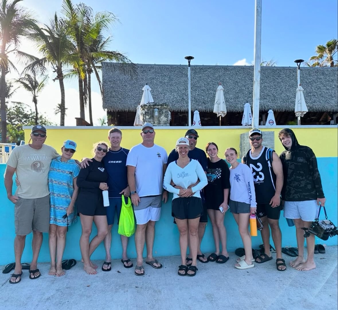 Group photo of eleven people at a beach location, smiling against a colorful wall and palm trees, indicating a fun outing or vacation.